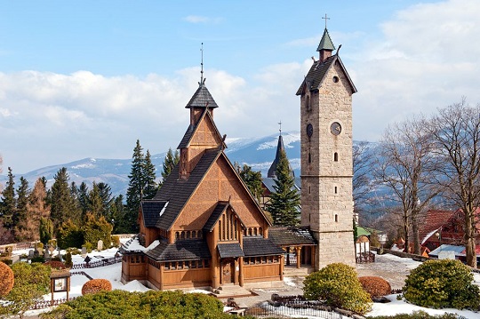 Igreja de madeira com torre de pedra à direita, montanhas ao fundo
