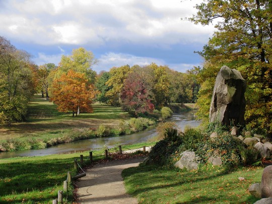 Imagem de uma parte do parque com rio, uma estátua e árvores no outono