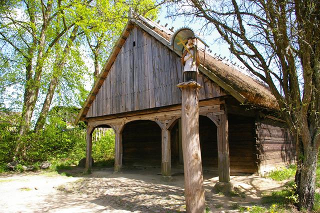 Pequeña iglesia de madera de carácter rural y construcción sencilla. Delante, la figura de un santo en una columna