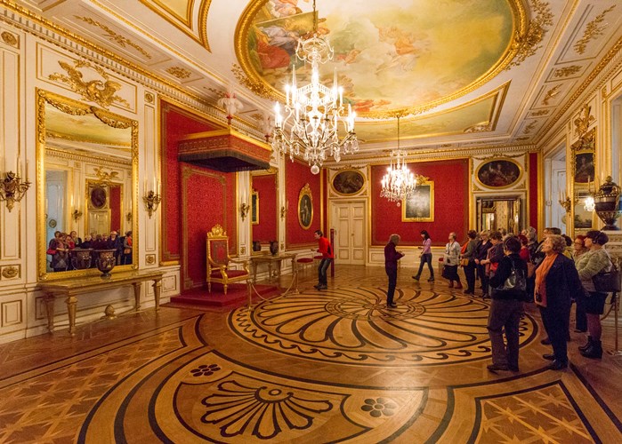 Sala de trono del castillo Real de Varsovia; las paredes en tono rojo o decorados con grandes espejos; suelo de parque conformado por tablillas de varias formas y tonos de madera que forman un motivo decorativo a la altura del trono
