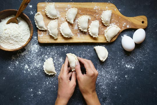 Empanadillas ya formadas sobre una tabla de madera y una que está siendo formada a mano; ingredientes básicos al lado: cuenco con harina y huevos
