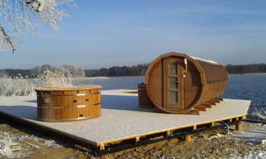 Sauna de madera y sauna en forma de barril sobre una tarina de madera, a orillas de un lago, rodeado de paisaje de invierno