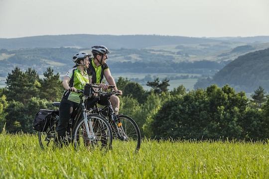 Foto di due persone in bici che si fermano ad ammirare il panorama. Sullo sfondo, delle montagne