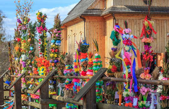 Palme pasquali create con fiori e nastri colorati, una chiesa in legno in sottofondo