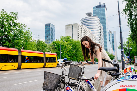 Una donna parcheggia la sua bici. Sullo sfondo i palazzi della città con un tram che passa.
